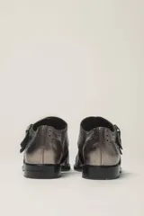 Zapato bajo de cuero negro, estilo monk strap con dos hebillas ajustables sobre el empeine. Presenta detalles de brogue (perforaciones) en la puntera y alrededor del borde superior. Tiene un taco bajo y ancho.