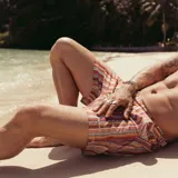 Short de baño con estampado de rayas horizontales y motivos geométricos en tonos naranja, marrón y blanco. Cuenta con pretina elástica y cordón ajustable.