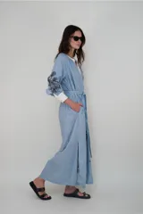 Vestido largo celeste con rayas verticales blancas, cuello mao con escote en V y lazo en la cintura. Presenta bordados de aves y palmeras en negro.