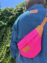 Riñonera fucsia de lona con correa regulable y detalles en cuero vacuno color marrón. Cuenta con bolsillo interno con cierre y bolsillo externo en la parte trasera.