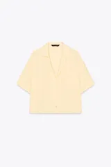 Camisa color amarillo pálido de tejido fluido, con cuello solapa, escote pico, manga corta, bolsillo de parche en el pecho y cierre frontal con botones.
