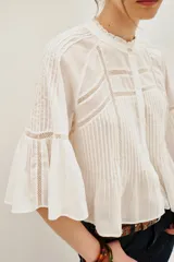 Camisa blanca de algodón con cuello alto y volados, mangas largas abullonadas con puños elásticos. Presenta detalles de encaje y frunces en el frente y la espalda.