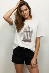 Remera de algodón blanca de manga corta y corte holgado, con cuello a la base. Presenta un estampado frontal en tono marrón oscuro de una jaula de pájaros con detalles de strass.