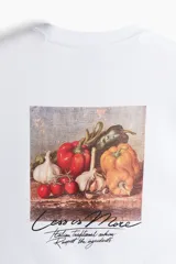 Remera blanca de algodón con cuello redondo y hombros caídos. Tiene un corte holgado y un pequeño estampado con la frase "Less is More" en el lado izquierdo del pecho.