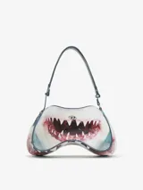 Bolso de hombro con forma estructurada y asimétrica, estampado con la boca abierta de un tiburón en tonos blancos, rojos y azules. Posee un asa ajustable y un pequeño logo metálico frontal.