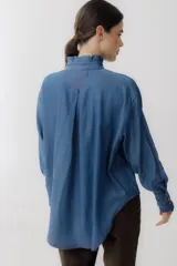 Camisa celeste de jean con cuello alto con volados y mangas largas abullonadas con puños con volados. Presenta botones de madera y costuras rojas a contraste.