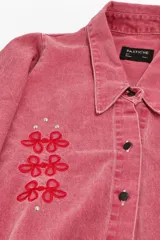 Camisa de jean color beige, de corte oversized, con cuello camisero y escote en V. Tiene mangas abullonadas, bolsillos delanteros, cierre con botones metálicos y bordado de flores en la parte delantera.