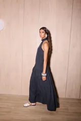 Vestido largo negro, holgado, con escote halter y espalda baja. Presenta un detalle de volado en el cuello y un maxi volado en la cadera. El material es tela Cupro, con apariencia similar a la gamuza.