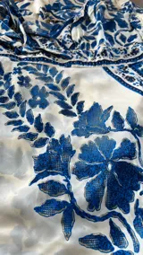 Vestido largo de verano, color blanco con estampado floral en azul índigo. Presenta tirantes finos, escote en V cruzado y un cinturón fino del mismo tejido que marca la cintura. La falda es amplia con volados.