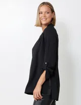 Blusa negra de manga larga con escote en V y pliegue central. La tela presenta un tejido piqué texturado sutil. Las mangas se pueden remangar y sujetar con una presilla y botón dorado.