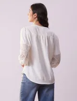 Blusa blanca de gasa con cuello a la base, escote en V y abertura central con botones. Tiene mangas 3/4 amplias con apliques de crochet y detalles plisados en la parte delantera.