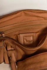 Bolso de mano tipo satchel o tote pequeño de cuero marrón con efecto lavado y apariencia vintage. Presenta dos asas superiores, múltiples cierres externos con tiradores de cuero y una etiqueta colgante metálica con logo.