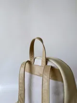 Mochila pequeña de cuero color gris claro y beige, con cierre superior y bolsillo frontal con cierre. Tiene asa de mano y correas ajustables para los hombros.