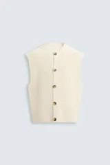 Chaleco color crema de punto con estructura, cuello redondo, sin mangas y cierre frontal con botones marrones.