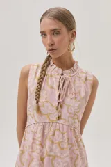 Vestido largo de algodón color rosa con estampado floral en tonos beige y dorado. Tiene cuello alto con lazo ajustable, corte imperio, falda escalonada con volados y diseño sin mangas.