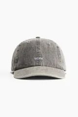 Gorra gris de algodón con diseño y ojales bordados en la parte superior. Tiene la palabra "WORK" bordada en el frente. Trabilla ajustable con hebilla de metal atrás y cinta antitranspirante de algodón.