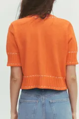 Chaqueta naranja de punto con cuello redondo y manga corta. Presenta bolsillos delanteros de parche con solapa y cierre frontal con botones. Detalle de pespuntes blancos a contraste.