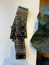 Cinto negro de cuero con hebilla metálica cuadrada y apliques de tachas plateadas y doradas.