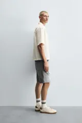 Bermuda de denim de algodón, color gris lavado, con corte relaxed fit y roturas visibles en la parte delantera de ambas piernas. Presenta cinco bolsillos y cierre frontal con cremallera y botón.