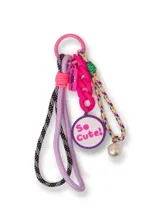Llavero con aro metálico rosado, cordones trenzados en blanco y negro y lila, cadena plástica rosada, dije redondo con la frase 'So Cute!' y una perla plástica.