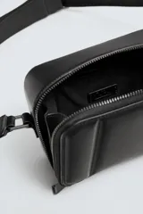 Bandolera negra acolchada con correa ajustable. Tiene un compartimento principal con cierre de cremallera, un bolsillo plano interior y tres compartimentos para tarjetas.