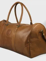 Bolso de viaje tipo duffel color marrón cuero, con doble asa corta y correa larga desmontable. Presenta un logo circular grabado en el centro y cierre superior con cremallera.
