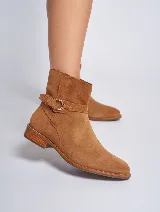 Bota corta de gamuza color marrón, con taco cuadrado y punta fina. Presenta una correa con hebilla dorada alrededor del tobillo y cierre lateral.