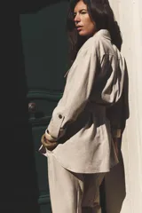Camisa color marrón con cuello solapa y manga larga acabada en vuelta con trabilla y botón. Bolsillos delanteros de plastrón con solapa. Bajo con aberturas laterales. Cierre frontal con botones y cinturón de la misma tela.