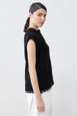 Chaleco negro de tejido de punto con cuello alto, cierre parcial con botones y detalle de pespunte blanco en el ruedo y el cuello. Sin mangas.