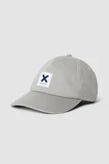 Gorra de béisbol color gris claro, con visera curva y parche frontal cuadrado con una 'X' azul marino y el texto 'blue banana' debajo.