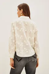 Camisa blanca de algodón con bordado floral en relieve, cuello clásico, cierre de botones frontales y mangas largas con puños fruncidos.