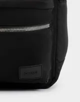 Mochila negra de tela deportiva con bolsillo frontal con cierre y tirador de cordón y goma. Tiene correas regulables, manija superior, doble cierre superior y etiqueta de goma.
