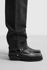 Bota negra de cuero con caña corta, horma cuadrada, suela a tono con ligero tacón y arnés decorativo a la altura del tobillo con detalles metálicos.
