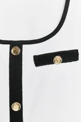 Camiseta blanca de corte slim fit, con cuello redondo y mangas cortas. Presenta detalles en negro en el cuello, los bordes de los falsos bolsillos y la botonadura frontal con botones dorados.