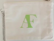 Sobre pequeño de tela color crudo con cierre superior y cadena metálica plateada. Presenta bordadas las iniciales AF en color verde.