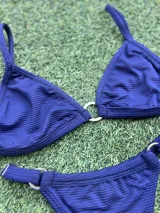 Bikini azul oscuro con tela texturada. El corpiño es triangular con una argolla de zamak en el centro y tirantes finos. La bombacha es colaless con argollas metálicas en los laterales.
