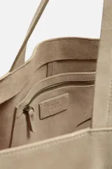Bolso tipo shopper color topo, fabricado en piel con acabado serraje. Cuenta con dos asas de mano y un bolsillo pequeño con cierre mediante cremallera en su interior.