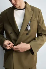 Blazer color verde oliva de corte regular, confeccionada en tejido con lana. Presenta cuello con solapas de pico, manga larga con puños abotonados, bolsillo de vivo en pecho, bolsillos de doble vivo en cadera, bolsillo interior y aberturas en la espalda. Cierre frontal cruzado con doble botonadura.
