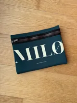 Clutch de neopreno color negro con cierre superior. Presenta estampado frontal con el texto "MILØ" en blanco y la dirección web "WWW.STOREMILO.COM" en la parte inferior.
