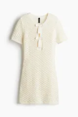 Vestido corto de tejido pointelle color crema, con cuello redondo, mangas cortas y abertura frontal con dos moños decorativos.