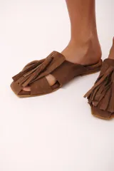 Zueco de cuero color marrón, estilo mule, con puntera cuadrada y tira ancha adornada con flecos. Suela plana.