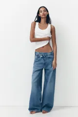 Pantalón de jean celeste de tiro alto, corte wide leg y largo hasta el tobillo. Presenta un diseño asimétrico en la cintura con cierre frontal cruzado con cremallera y botones metálicos.