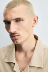 Camisa de denim de algodón, color beige claro, con corte relajado, cuello bowling y manga corta. Presenta cierre frontal con botones a presión y un pequeño bordado a tono en el pecho.