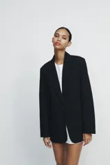 Blazer negra de corte clásico, con cuello de solapa y escote en pico. Tiene mangas largas con hombreras, bolsillos con solapa en el delantero y un bolsillo de vivo en el pecho. Está forrada y se cierra con un botón frontal.