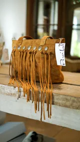 Bolso de cuero color beige con doble asa corta y correa larga extraíble. Presenta detalle de flecos largos en la parte frontal y herrajes metálicos plateados. Interior forrado con bolsillos. Marca Brava.