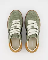 Championes Coolway modelo Goal, color verde musgo, confeccionados en serraje suave con detalles en contraste de color. Cuentan con lengüeta preformada de piel acolchada, forro interior en piel, cordones planos de algodón bicolor intercambiables, cuello ligeramente acolchado, puntera reforzada con pespuntes decorativos y suela flexible de goma y caucho.