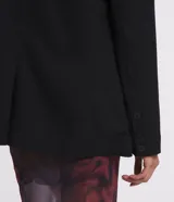 Blazer negro de sastrería con corte alargado, cuello con solapa, manga larga, botón delantero, bolsillos fake y cadena decorativa en la solapa.