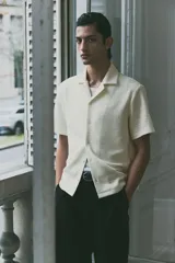 Camisa de manga corta color blanco hueso con textura. Tiene cuello abierto, tapeta lisa y bajo recto.