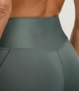 Conjunto deportivo femenino compuesto por un top deportivo de tirantes anchos y un legging de cintura alta. El conjunto es de color verde oscuro o verde oliva, con un acabado liso y ajustado al cuerpo. El legging presenta un detalle de costura o sesgo aplicado sobrepuesto en los laterales.