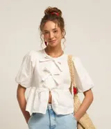 Blusa blanca de tricolina con cuello redondo, manga corta abullonada, cierre frontal con lazos y volado en la cintura.